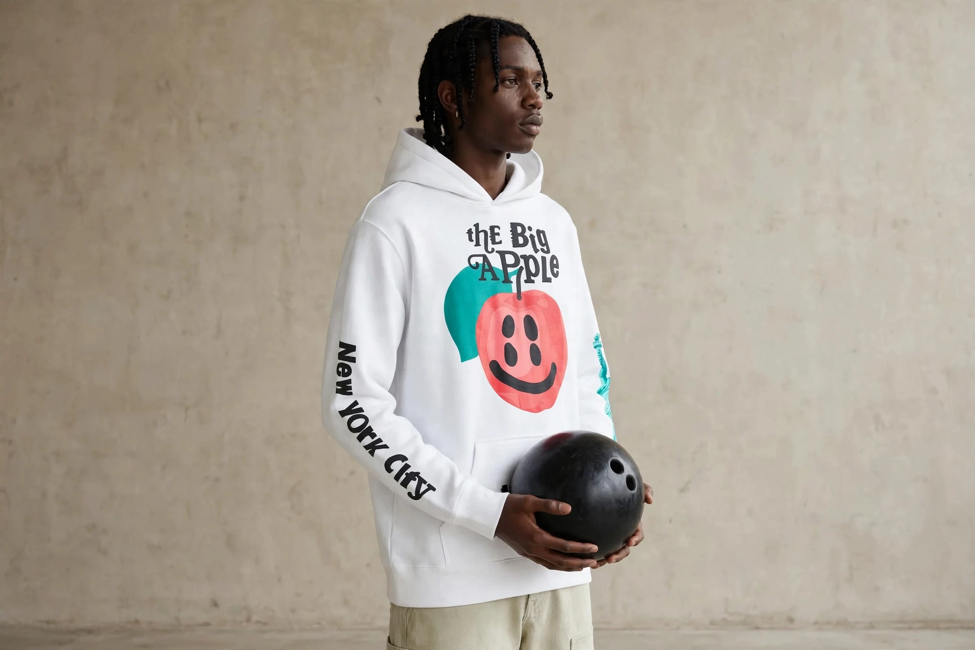 Male model wearing white Big Apple hoodie with red apple smiley and NYC graphics preserved exactly, holding black bowling ball at waist, beige concrete studio background, 1920 by 1280 landscape frame, premium editorial